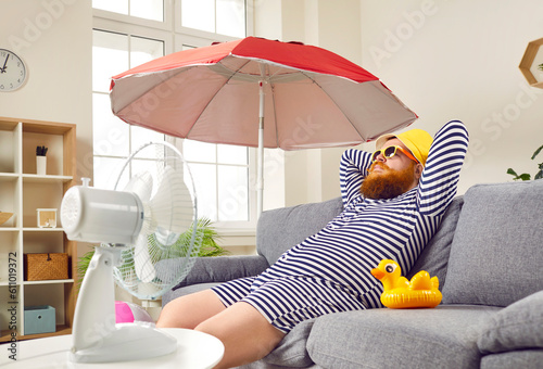 Obraz na plátně Funny man in sunglasses and bright yellow panama hat is sitting with rubber duck on the sofa in the living room under umbrella, imagining, dreaming of vacation at sea, blown by the wind from the fan