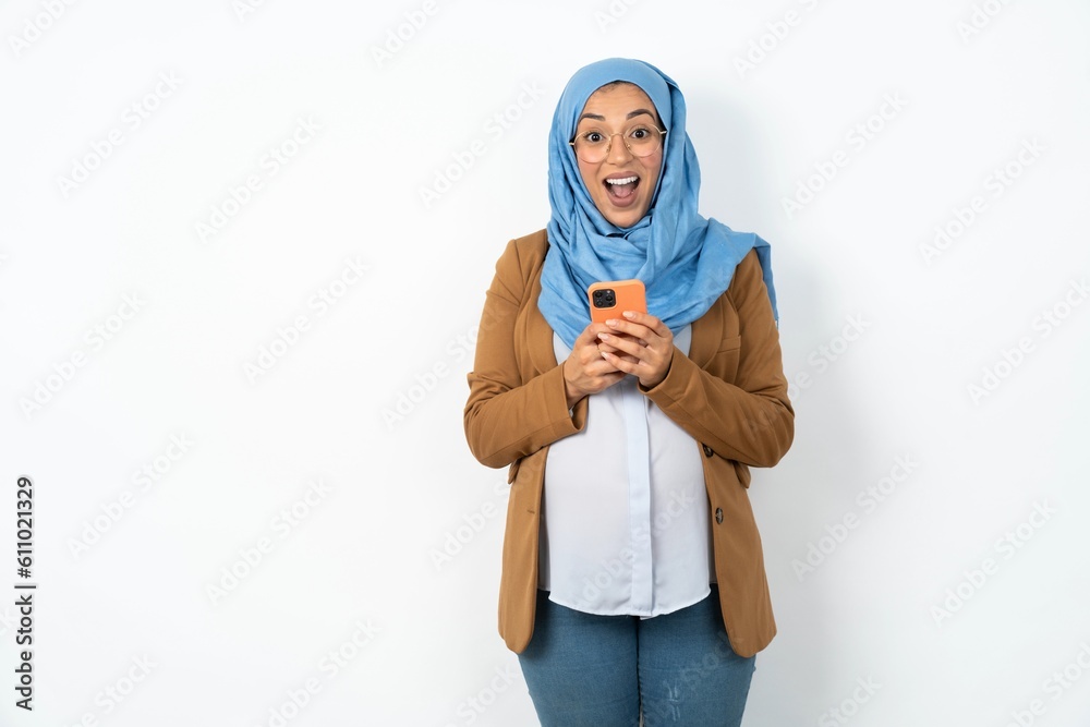 Excited Young beautiful pregnant muslim woman wearing hijab over white background holding smartphone and looking amazed to the camera after receiving good news. Excited Young beautiful pregnant muslim woman wearing hijab over white background holding smartphone and looking amazed to the camera after receiving good news.