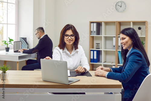 Loan broker, financial advisor, business manager, or real estate agent meeting with client. Two happy beautiful women sitting at office desk, looking at laptop PC and discussing different options