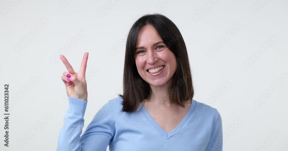 A Caucasian woman stands on a white background, flashing a bright smile as she shows a victory sign with her hand. The V sign is a gesture of triumph or peace. A Caucasian woman stands on a white background, flashing a bright smile as she shows a victory sign with her hand. The V sign is a gesture of triumph or peace.
