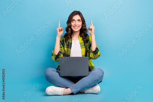Full length portrait of cheerful pretty person sit floor use netbook look direct fingers up empty space isolated on blue color background © deagreez