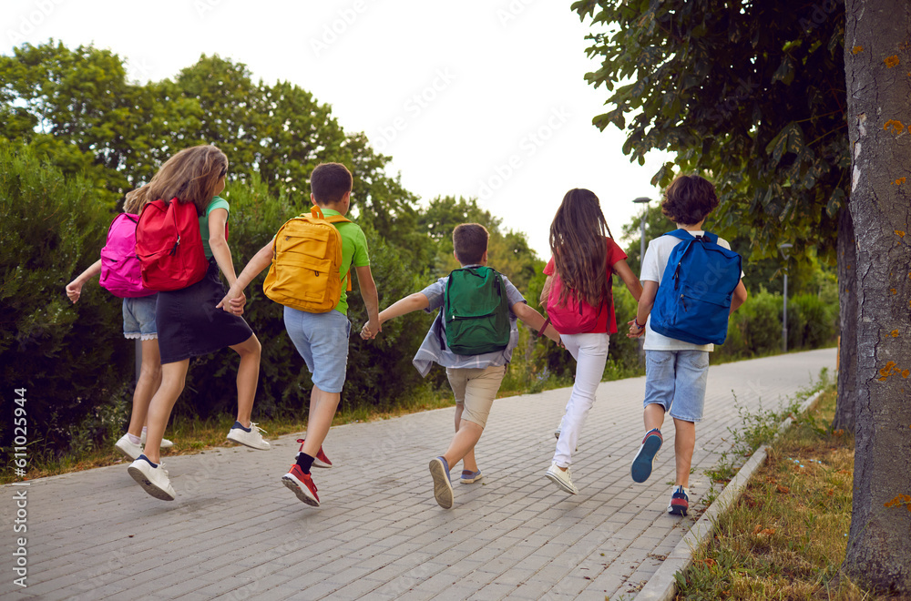 Little best school friends with backpacks on their shoulders all run on sidewalk in park. Rear view of children with colored backpacks running holding hands. Concept education, children and learning.