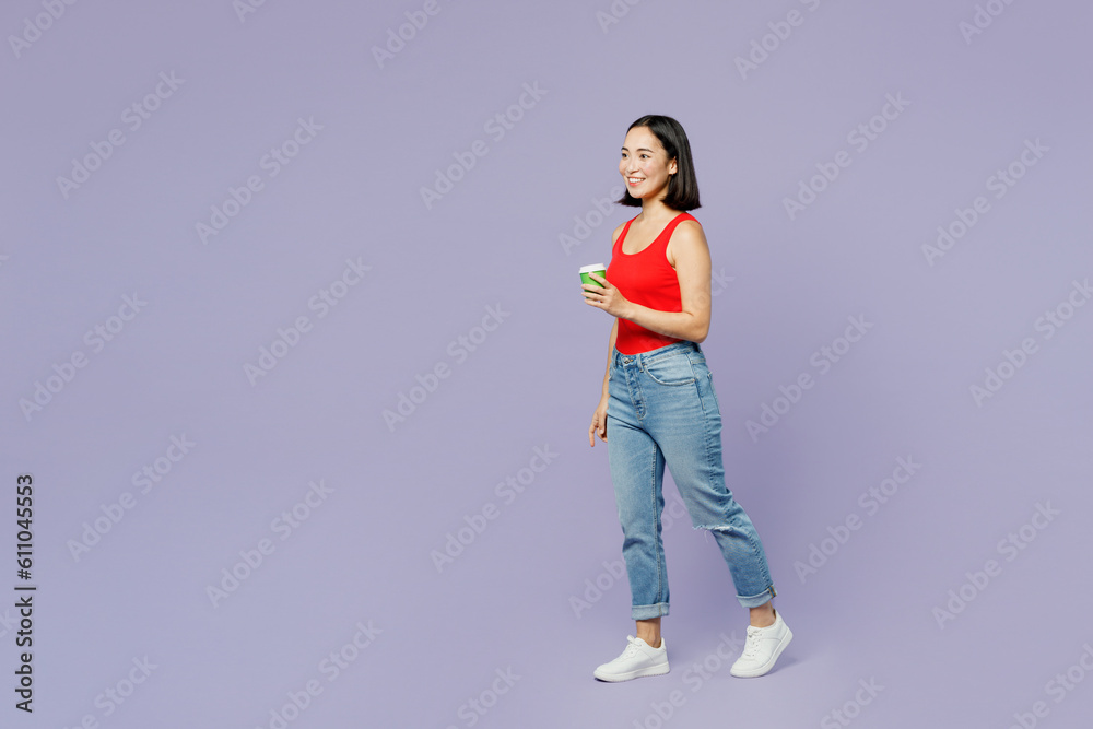 Full body young woman of Asian ethnicity wearing casual clothes red tank shirt hold takeaway delivery craft paper brown cup coffee to go walk isolated on plain pastel light purple background studio. Full body young woman of Asian ethnicity wearing casual clothes red tank shirt hold takeaway delivery craft paper brown cup coffee to go walk isolated on plain pastel light purple background studio.