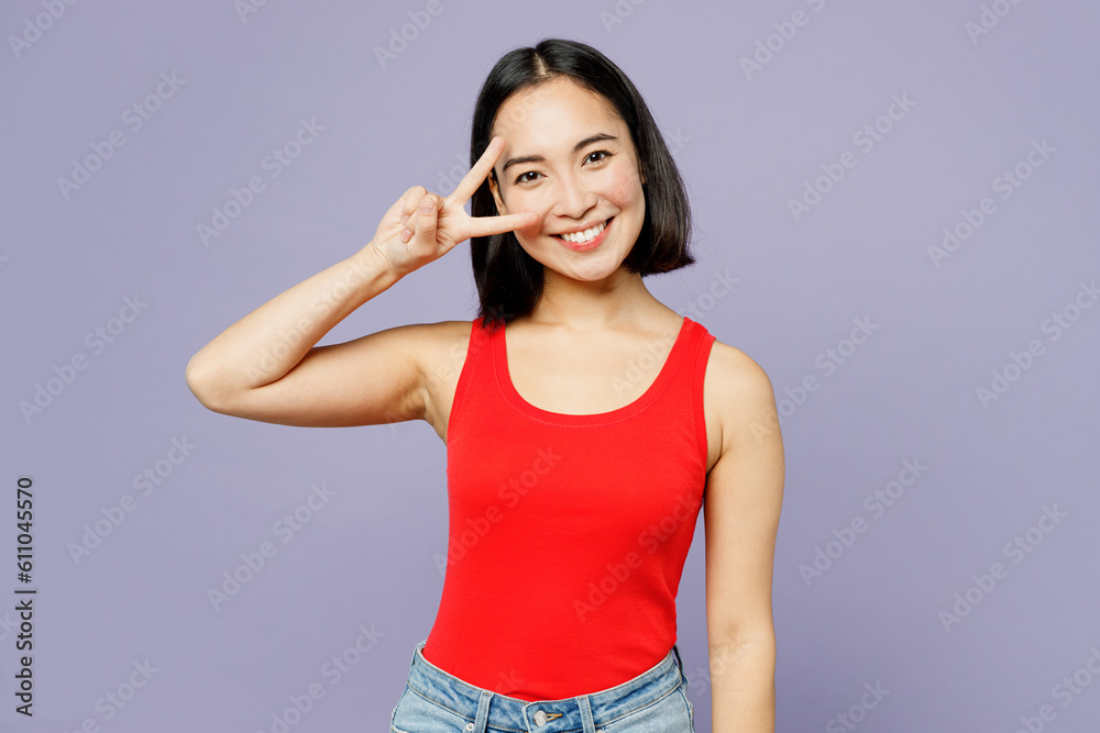 Young happy woman of Asian ethnicity she wear casual clothes red tank shirt look camera show cover eye with v-sign isolated on plain pastel light purple background studio portrait. Lifestyle concept. Young happy woman of Asian ethnicity she wear casual clothes red tank shirt look camera show cover eye with v-sign isolated on plain pastel light purple background studio portrait. Lifestyle concept.