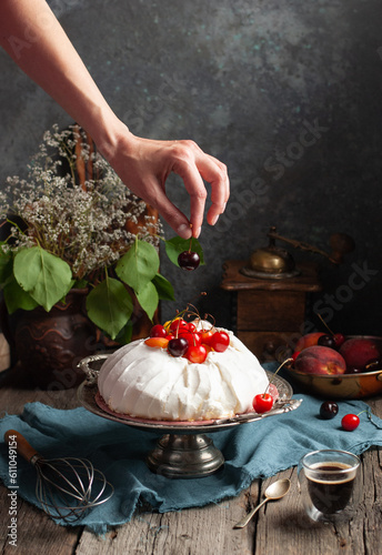 a woman's hand puts a cherry on a meringue homemade cake with sweet cherries and chocolate syrup on a dark background surrounded by vintage crockery and flowers