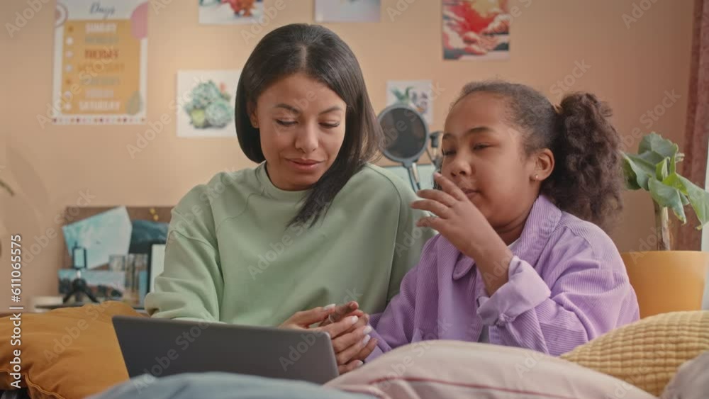 Waist up of young Biracial woman and her pretty 10 year old daughter chatting while sitting by laptop on bed in vibrant kids bedroom Waist up of young Biracial woman and her pretty 10 year old daughter chatting while sitting by laptop on bed in vibrant kids bedroom