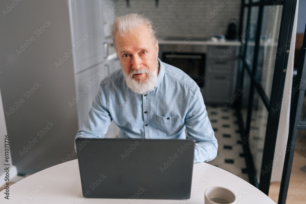 Portrait of handsome mature man using laptop sitting at table with coffee cup in modern kitchen room. Confident senior male adult posing at home working on pc, looking at camera, typing on keyboard. Portrait of handsome mature man using laptop sitting at table with coffee cup in modern kitchen room. Confident senior male adult posing at home working on pc, looking at camera, typing on keyboard.