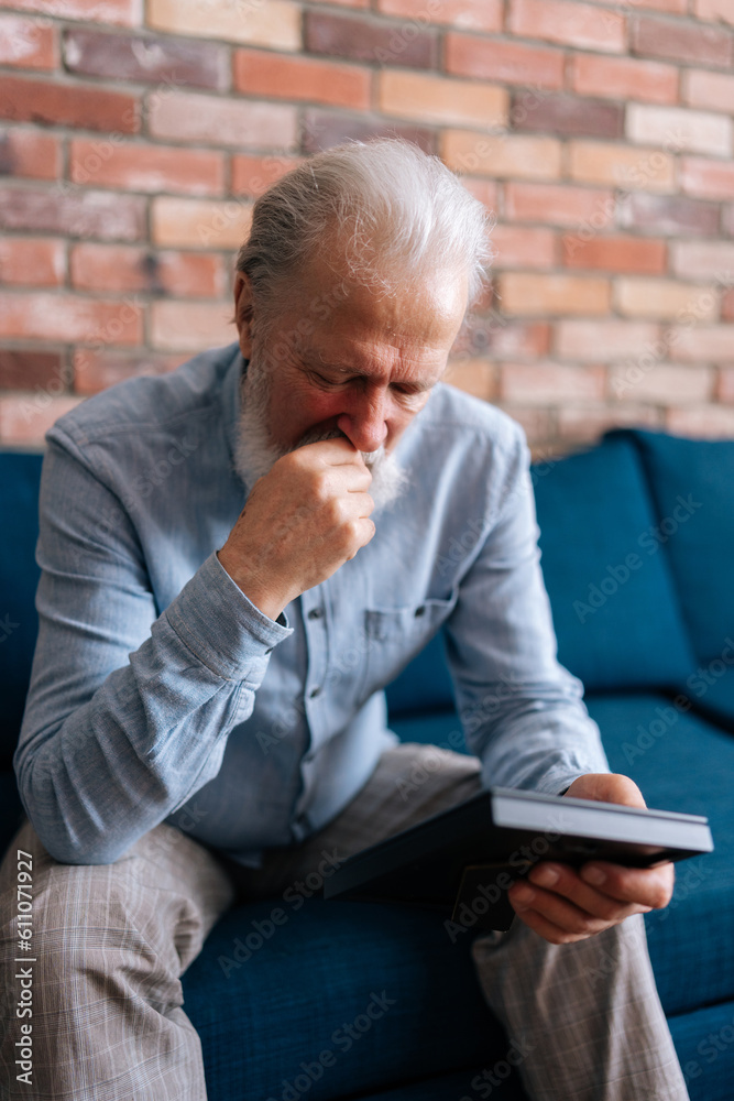 Vertical portrait of depressed elderly bearded man sitting on sofa at home, looking at picture of wife in frame and crying. Concept of nostalgia, grief, longing, loneliness in old age. Vertical portrait of depressed elderly bearded man sitting on sofa at home, looking at picture of wife in frame and crying. Concept of nostalgia, grief, longing, loneliness in old age.