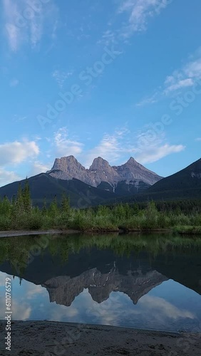 Timelapse sunset video of Three Sisters view seen in Canmore outside of Banff National Park with stunning reflection in water below. Clouds moving across sky with colours, blue sky, mountain peaks.