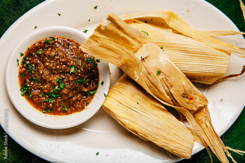 A platter of cooked wrapped tamales and a bowl of peppery salsa on a green table, closeup overhead photo