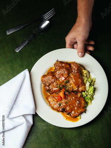 A hand serves cheese and pork schnitzel covered in Madeira sauce and tomatoes atop salad on a white platter and green table