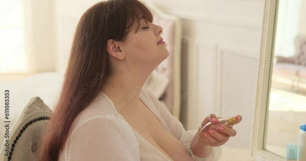 Confident plus-size Caucasian woman sits by her vanity table in the serene bedroom. With a gentle smile, she gracefully sprays her favorite perfume, letting its alluring fragrance uplift her spirits. Confident plus-size Caucasian woman sits by her vanity table in the serene bedroom. With a gentle smile, she gracefully sprays her favorite perfume, letting its alluring fragrance uplift her spirits.