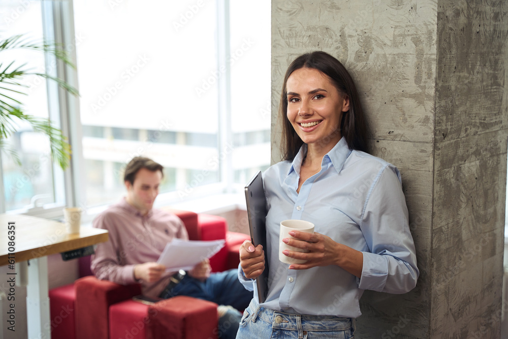 Joyful company employee with teacup and laptop leaning against pillar Joyful company employee with teacup and laptop leaning against pillar