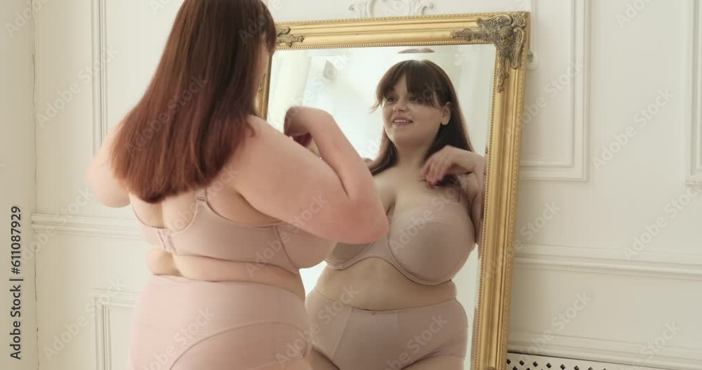 A plus-size Caucasian woman stands by the mirror in her bedroom, beaming with a smile. In this uplifting moment, she embraces her beauty, radiating confidence and self-love.