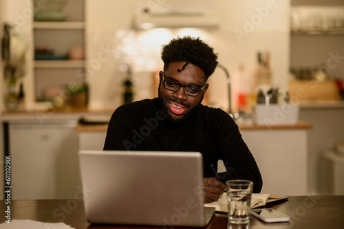 An interracial hipster is sitting at home at night and following an online course on a laptop.