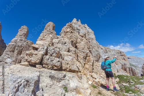 Young woman with long air and cap taking photos with phone of dolomite peaks. Scenic landscape on the nature, Mount Latemar, Trentino, Italy. Traveling photography and outdoor sport activity concept