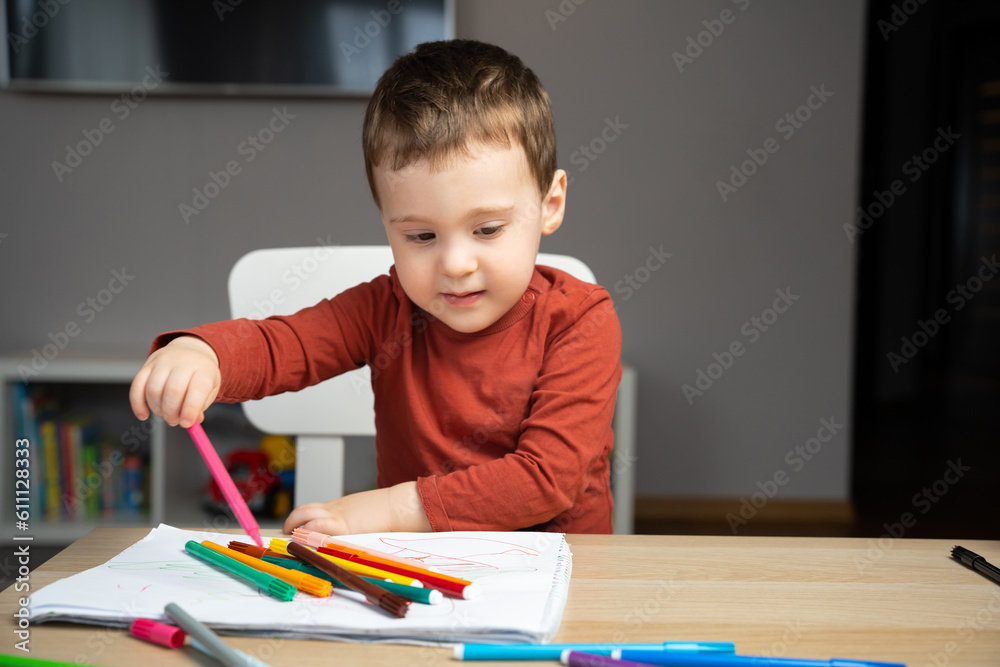 A cute happy little toddler boy of two years old draws with markers in the album in the children's room at home. Educational activities for kids. Selective focus A cute happy little toddler boy of two years old draws with markers in the album in the children's room at home. Educational activities for kids. Selective focus