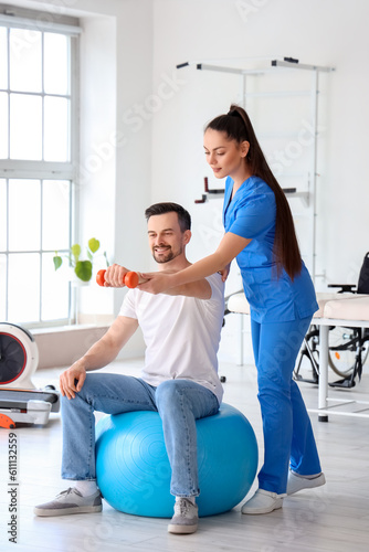 Foto Young man training with dumbbell and physiotherapist in rehabilitation center