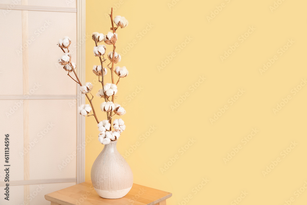 Vase of cotton sprigs on wooden stool with dressing screen near yellow wall