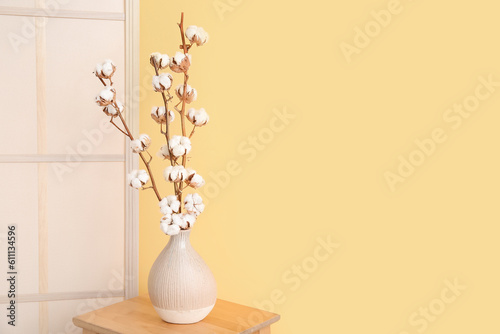 Vase of cotton sprigs on wooden stool with dressing screen near yellow wall