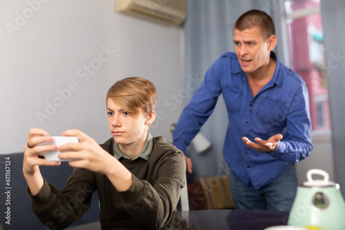 Teenage school boy sitting at table at home interior with smartphone and playing game, father trying to speak with him