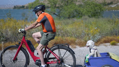 Elderly senior man biking on an e-bike on a trail pulling a trailer with a dog in it.