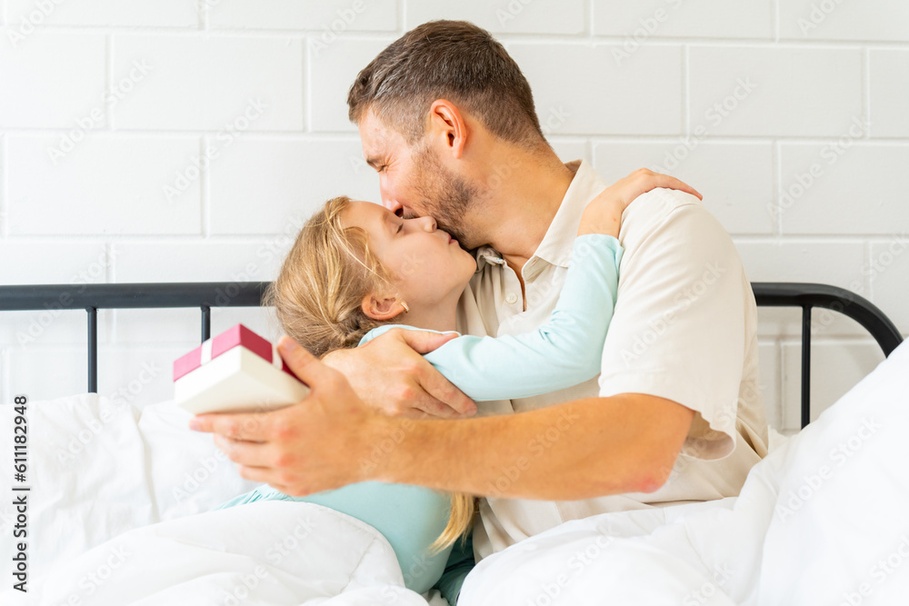 Fototapeta premium Father wake up a sleeping child daughter on the bed in bedroom in sunny morning. Happy family start a new day together with freshness. Little girl waking up with smiling.