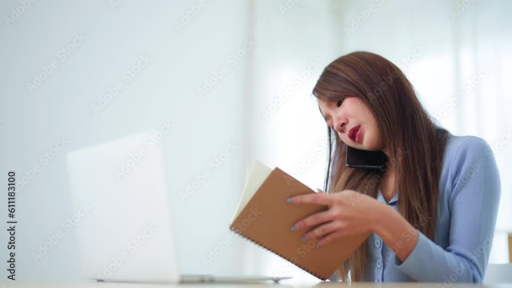 Young asian woman talking mobile smartphone while working on computer laptop on desk in living room at home Young asian woman talking mobile smartphone while working on computer laptop on desk in living room at home