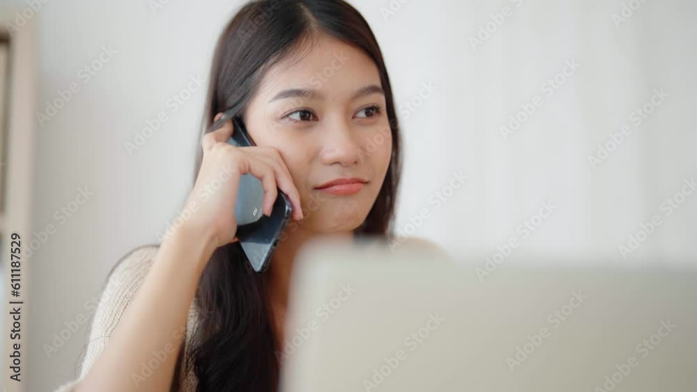 Young asian woman talking mobile smartphone while working on computer laptop on desk in living room at home