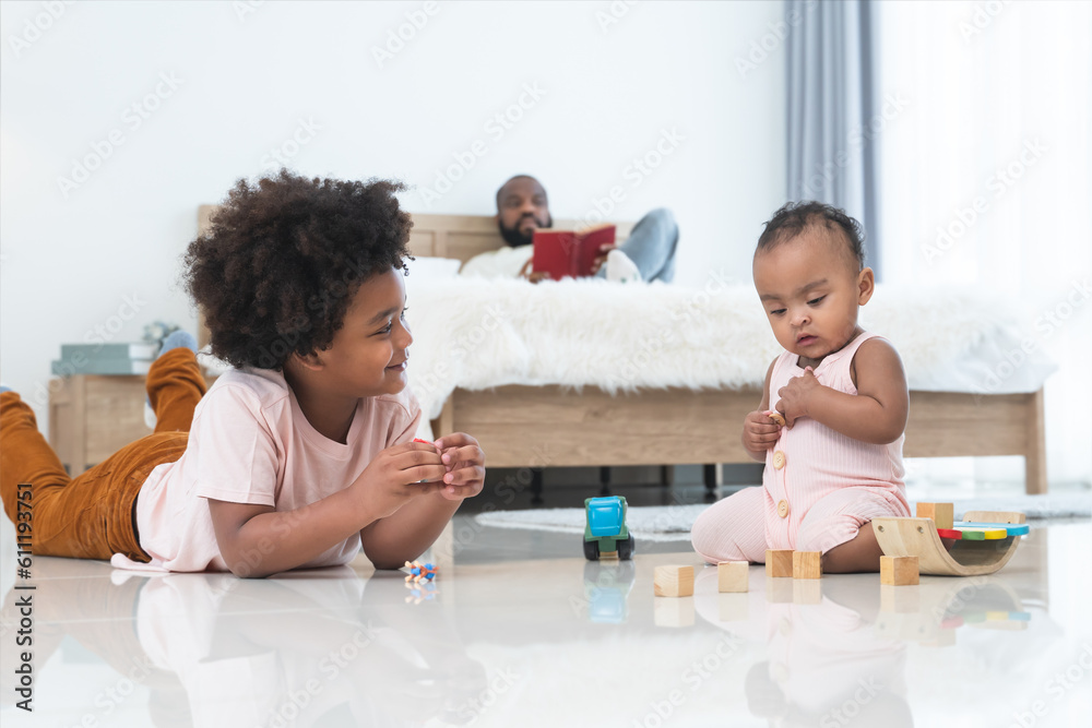 African family, little boy and cute toddler baby girl, brother and sister have fun playing model toys and wooden blocks together on floor while single dad lying on bed, reading book, looking care kids African family, little boy and cute toddler baby girl, brother and sister have fun playing model toys and wooden blocks together on floor while single dad lying on bed, reading book, looking care kids