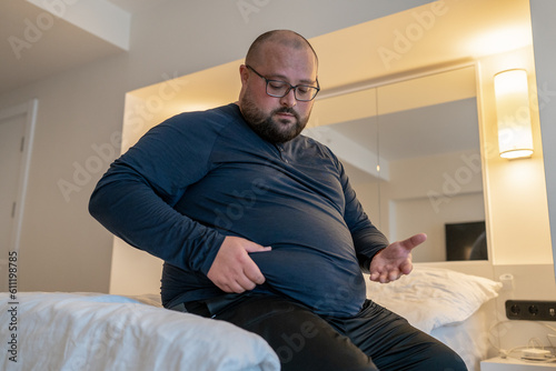 Huge man suffering from extra weight touching stomach sitting on bed in bedroom. Overweight bearded balded middle aged male with unhealthy body noticed problem. Weight control, overeating concept.