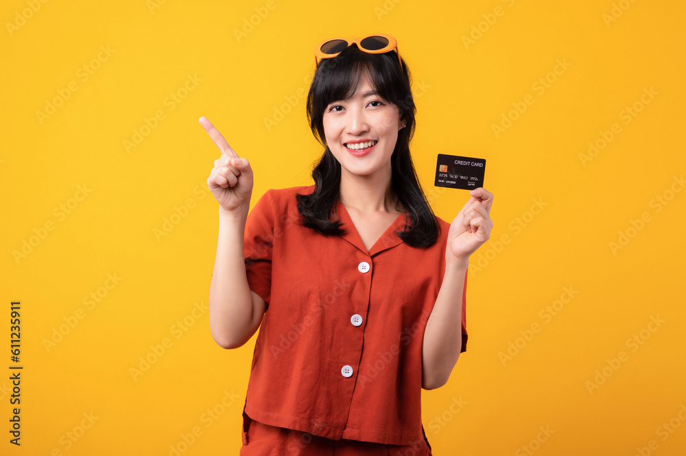 Portrait young beautiful asian woman happy smile dressed in orange clothes showing credit card and point finger to free space isolated on yellow background. Pay and purchase shopping payment concept. Portrait young beautiful asian woman happy smile dressed in orange clothes showing credit card and point finger to free space isolated on yellow background. Pay and purchase shopping payment concept.