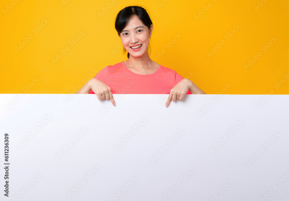 young asian sports fitness woman happy smile wearing pink sportswear standing behind the white blank banner or empty space advertisement board against yellow background. wellbeing lifestyle concept. young asian sports fitness woman happy smile wearing pink sportswear standing behind the white blank banner or empty space advertisement board against yellow background. wellbeing lifestyle concept.