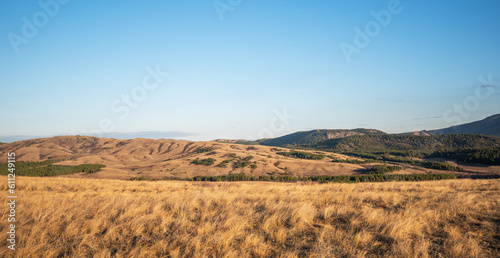 Colorful autumn view the picturesque valley. Forested slopes and mountains in the distance in a blue haze. Dry grass and forest on a sunny day under a blue cloud sky on a warm autumn day in October.