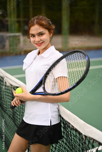 Portrait of smiling female tennis player with racket and ball standing by net at the outdoor tennis court © Prathankarnpap