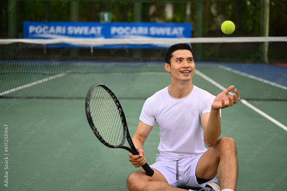 Happy sportive asian man with racket throwing tennis ball in air, resting on tennis court. Exercise, sports and wellness concept