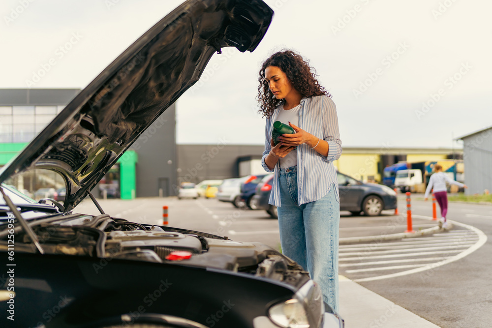 Obraz premium A young woman stands in front of a car with the hood up and reads the instructions on how to change the oil