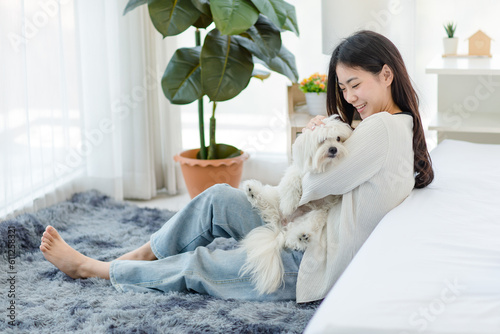 Asian young happy cheerful female owner sitting smiling on fluffy carpet floor holding hugging cuddling embracing best friend companion dog white long hair mutt shih tzu together in bedroom at home