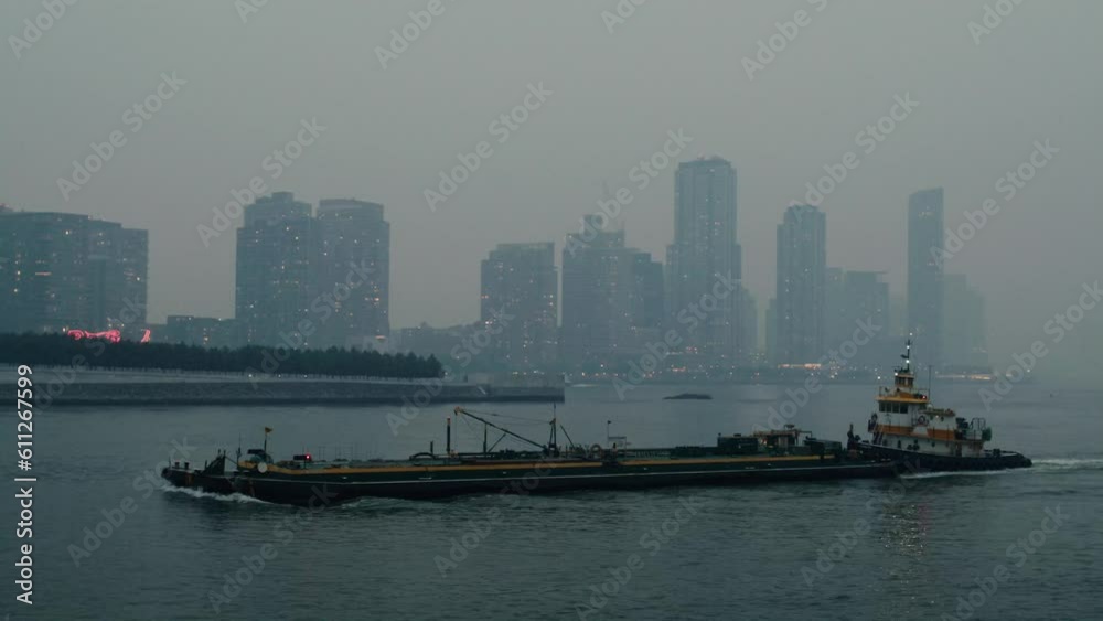 New York City covered in smoke from wildfires seen from across the east river. Tracking shot of tug boat and barge navigating along east river. slow motion 40fps. New York City covered in smoke from wildfires seen from across the east river. Tracking shot of tug boat and barge navigating along east river. slow motion 40fps.