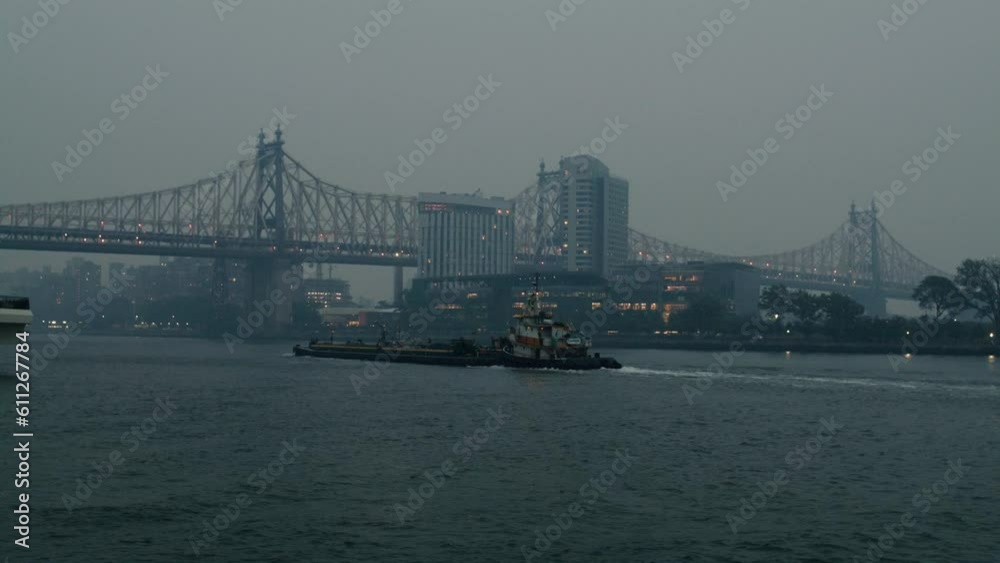 New York City covered in smoke from wildfires seen from across the east river. Tracking shot of tug boat and barge navigating along east river. slow motion 40fps. New York City covered in smoke from wildfires seen from across the east river. Tracking shot of tug boat and barge navigating along east river. slow motion 40fps.