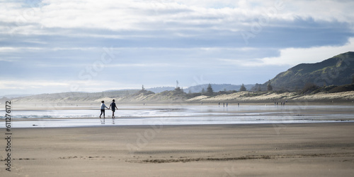 Couple holding hands and walking barefoot on Muriwai beach. Unrecognizable people walking in the distance. Auckland.