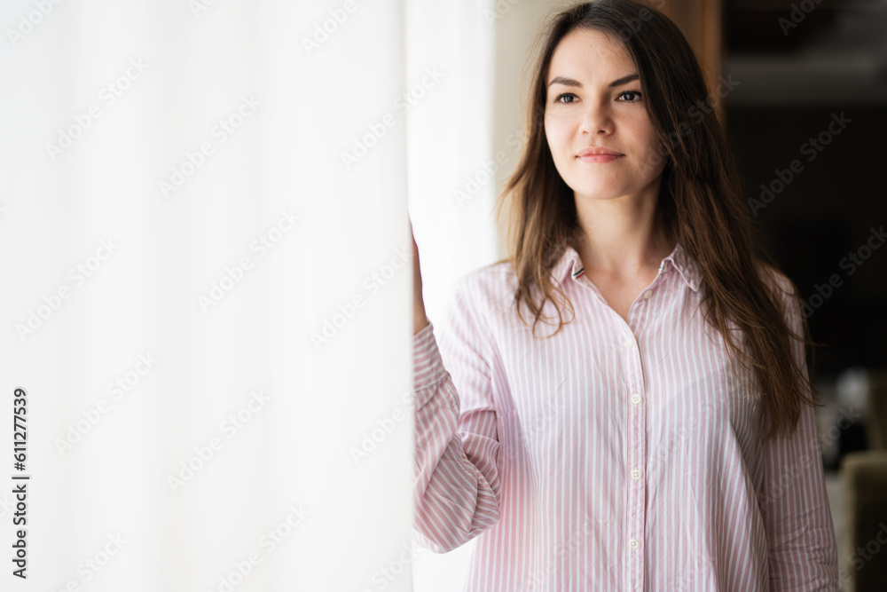 Young beautiful brunette woman at the window light background, touches the curtain with her fingers. Young beautiful brunette woman at the window light background, touches the curtain with her fingers.
