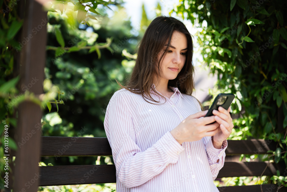 Young freelancer woman in shirt sit on bench in summer park outdoors rest and use mobile cell phone. Young freelancer woman in shirt sit on bench in summer park outdoors rest and use mobile cell phone.