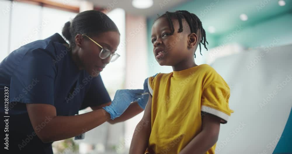 Young African American Boy Sitting In The Chair In Bright Hospital And Getting His Flu Vaccine. Female Black Nurse Is Performing Injection. Professional Woman High-Fives A Worried Kid For Being Brave. Young African American Boy Sitting In The Chair In Bright Hospital And Getting His Flu Vaccine. Female Black Nurse Is Performing Injection. Professional Woman High-Fives A Worried Kid For Being Brave.
