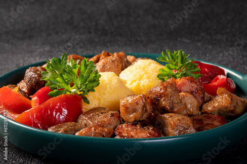 Romanian festive dish pomana porcului consisting of pieces of fried pork and sausages served with polenta and pickled red peppers close-up on a plate. Horizontal.