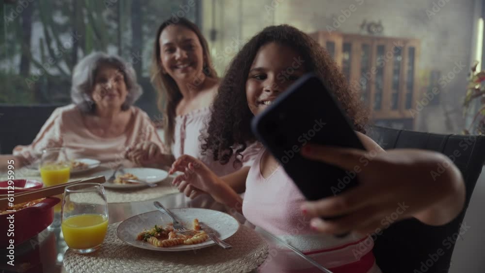 criança brasileira sentada na mesa de jantar fazendo uma selfie com sua mãe e sua avó durante o almoço de dia das mães