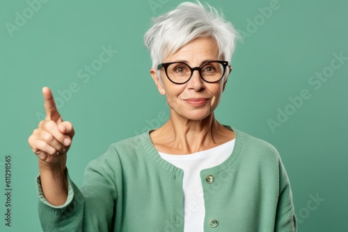 Close-up portrait photography of a glad mature woman making a i have an idea gesture with a finger up against a pastel green background. With generative AI technology