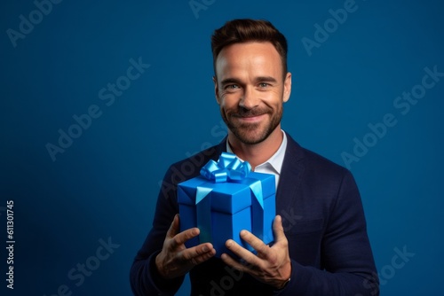 Headshot portrait photography of a glad boy in his 30s holding a gift against a sapphire blue background. With generative AI technology © Markus Schröder