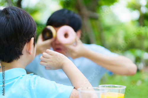 Happy cheerful Asian father and son eating a doughnut together during picnic in the garden. Excited lovely little boy eating a doughnut with his dad. Family weekend activity concept.