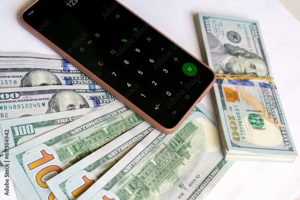 Fototapeta premium A stack of one hundred dollar bills and a calculator on a smartphone. A stack of one hundred dollar bills and a smartphone on a light background.